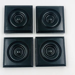 May include: Four square, black coasters with a concentric circle design in the center. The coasters have a raised border and a glossy finish. The coasters are arranged in a square formation.