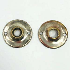 May include: Two antique metal door knob backplates. The backplates are circular with a central hole and two smaller holes for screws. One is silver-toned, the other is gold-toned, both showing signs of age and wear.