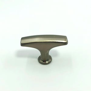 Puede incluir: Un tirador de armario de níquel cepillado con un diseño en forma de T. El tirador tiene una parte superior rectangular y una base redondeada, adecuado para cajones o puertas de armarios. El herraje es de metal y tiene un acabado mate.