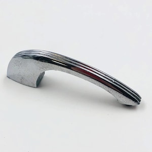 May include: A chrome-plated door handle with a curved, streamlined design. The handle features a series of parallel grooves along its length, giving it a textured appearance. The handle is likely for a vintage car or a retro-style door.