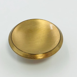May include: A round, brass-colored knob with a brushed metal finish. The knob has a slightly concave center and a raised, rounded edge. The overall shape is smooth and simple, suitable for furniture or decorative use.