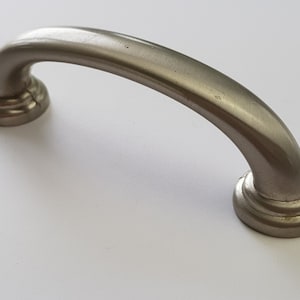 May include: A brushed nickel cabinet pull with a rounded, arched shape. The pull has a smooth, polished finish and a simple, modern design.
