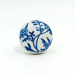 May include: A white ceramic cabinet knob with a blue floral design. The knob is round and features a detailed pattern of leaves and dotted clusters. The design is symmetrical and covers the entire surface of the knob.