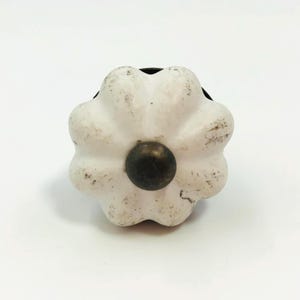 以下が含まれることがあります： 花のようなスカラップ形状をした白いセラミック製の家具用ノブで、使い古されたような仕上げが施されています。ノブの中央には暗い色の円形金属製アクセントパーツがあります。全体的に丸みを帯びたデザインで、6つの花びらのようなセクションに分かれています。