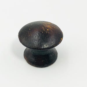 Puede incluir: Perilla de madera en forma de hongo, de color marrón oscuro, con una parte superior redondeada y una base cilíndrica. La perilla tiene un acabado envejecido con vetas de madera visibles y un aspecto ligeramente desgastado.