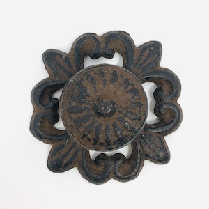 May include: A circular metal knob featuring a dark brown and black finish with a textured surface. The knob is centred on a decorative, openwork metal backplate that displays a symmetrical, scrolling floral pattern in matching dark tones.