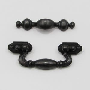 May include: Two antique-style drawer pulls in a dark, matte finish. One is a simple bar pull, and the other is a drop pull with curved arms and decorative end pieces. Both are made of metal and are likely for furniture.