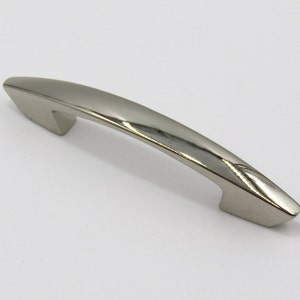 Pode incluir: Uma puxador de armário arqueado de cor prateada com um acabamento liso e polido. O puxador tem uma forma ligeiramente curva e uma ponta pontiaguda em cada lado.