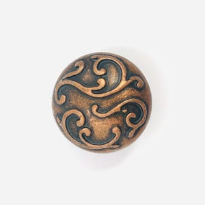 May include: A round cabinet knob featuring a metallic copper finish with a dark, recessed scrollwork pattern. The surface displays a raised, swirling design that contrasts with the darkened background, creating a textured appearance on the circular face.
