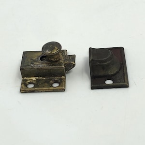 May include: An antique brass-toned latch and strike plate set. The latch features a rounded knob and a rectangular body with two screw holes. The strike plate is a matching dark brass color and has a curved design with a single screw hole.