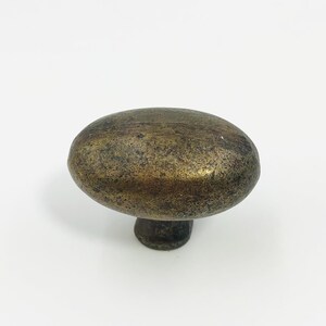 May include: An oval-shaped cabinet knob with a dark, weathered brass finish. The surface features a mottled, aged texture with visible dark brown and gold-toned patina throughout the metal, supported by a small, cylindrical base attachment.