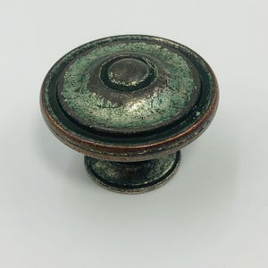 Könnte beinhalten: Ein verwitterter Schrankknauf mit einem antiken Finish. Der Knauf hat ein kreisförmiges Design mit einem erhöhten Mittelknopf und einem dekorativen Rand. Die Farbgebung umfasst Bronze-, Grün- und Silbertöne, was ihm ein antikes Aussehen verleiht.