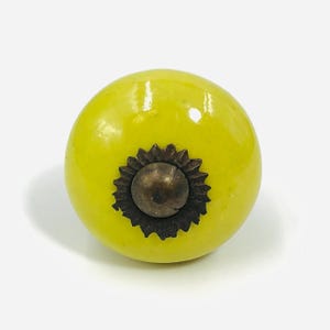 May include: A round, bright yellow ceramic cabinet knob with a smooth, glossy finish. The center features a dark, metallic, sunburst-shaped decorative plate surrounding a small, raised, circular metal dome in a weathered bronze tone.