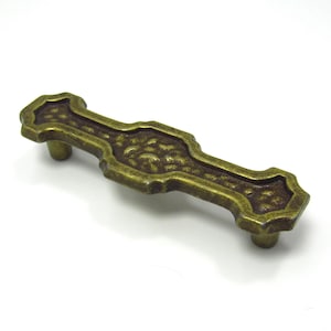 May include: A vintage-style brass drawer pull with a textured center and decorative edges. The hardware features two cylindrical posts for mounting and is approximately 5 inches long.