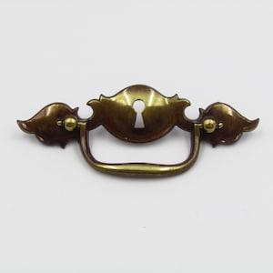 May include: A brass cabinet pull with a keyhole design and ornate wings. The pull has a brown patina finish.