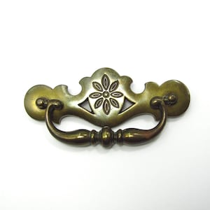 May include: Antique brass drawer pull with a floral design in the center. The pull has a curved handle and decorative end pieces. This vintage hardware is ideal for furniture restoration projects.