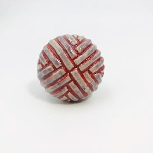 May include: A round ceramic knob featuring a textured, woven-like pattern of raised, light lavender-grey bars separated by deep, glossy red grooves. The surface has a smooth, glazed finish, creating a distinct contrast between the colors.