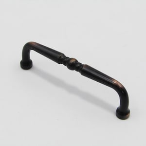 Peut inclure: Une poignée de meuble en bronze foncé avec une poignée incurvée et un centre décoratif. La quincaillerie présente un design classique avec des extrémités arrondies et une section centrale détaillée.