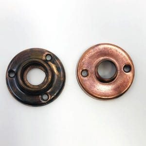 Könnte beinhalten: Zwei antike Türrosetten in Kupferfarbe. Die Rosetten sind rund mit einem zentralen Loch und drei kleineren Löchern für Schrauben. Eine Rosette hat eine dunklere, gealterte Patina, die andere ist kupferfarbener.