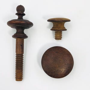 Puede incluir: Tres piezas de herrajes de muebles de madera antiguos. Las piezas incluyen un tornillo grande y ornamentado con una parte superior decorativa, un tornillo más pequeño con una parte superior simple y un disco de madera redondo y plano. La madera tiene un tono marrón cálido.