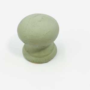 Puede incluir: Un pomo de madera redondo verde salvia con base de pedestal. El pomo tiene una superficie ligeramente texturizada y está sobre un fondo blanco. Esta ferretería decorativa es adecuada para muebles y armarios.