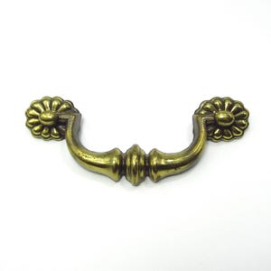 May include: A vintage-style brass drawer pull with a floral design on each end. The handle curves gracefully between the floral accents, and the brass has a slightly aged appearance. This hardware is suitable for cabinets and drawers.