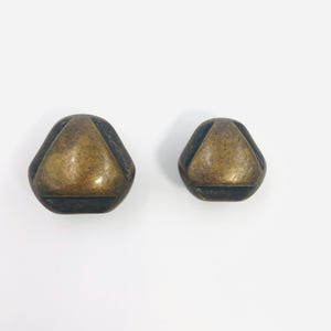 May include: Two brass-toned metal buttons with a rounded triangular shape. Each button features a smooth, raised central triangle surrounded by a recessed, dark-toned border, creating a distinct contrast in texture and depth on the surface.