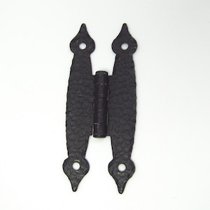 Puede incluir: Bisagra de hierro forjado negro con superficie texturizada y extremos puntiagudos decorativos. La bisagra tiene un barril central para el movimiento, diseñada para puertas o portones. El herraje se muestra sobre un fondo blanco.