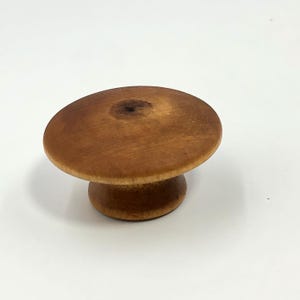 Puede incluir: Un pequeño pomo de madera redondo con forma de hongo. El pomo tiene una parte superior plana y circular y una base redondeada, todo en un tono de madera natural y cálido. El pomo es probablemente para un cajón o un armario.