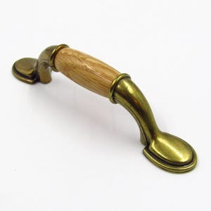 Puede incluir: Un tirador de armario con un centro de madera marrón claro y acabado de latón antiguo. El tirador tiene un diseño curvo con extremos decorativos, adecuado para muebles o armarios. El herraje está diseñado para una fácil instalación.