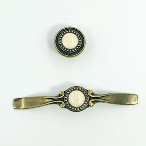 May include: Two antique brass cabinet hardware pieces. One is a round knob with a cream-colored center and a dark border. The other is a matching pull with a similar cream center and decorative detailing.
