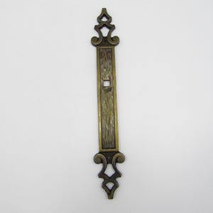 May include: An antique brass-toned cabinet pull with an ornate design. The pull features a rectangular center with a textured surface and decorative scrollwork at the top and bottom. The hardware is approximately 7 inches long.