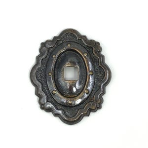 May include: Ornate, antique-style metal escutcheon plate with a dark bronze finish. The oval-shaped plate features a central square opening and is embellished with decorative rivets and a scalloped edge. A vintage hardware element.