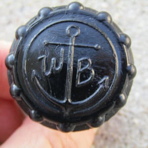 以下が含まれることがあります： 黒い丸いノブで、中央に錨のデザインと「WB」の文字があります。ノブはテクスチャ加工された表面とビーズの縁があります。デザインは浮き彫りになっており、文字は様式化されたフォントです。航海をテーマにしています。