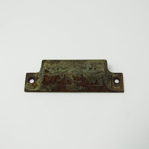 May include: A weathered, rectangular metal bracket with two circular holes on either end. The bracket has a raised central section and a mottled, aged appearance, suggesting exposure to the elements. The color is a mix of brown and green.