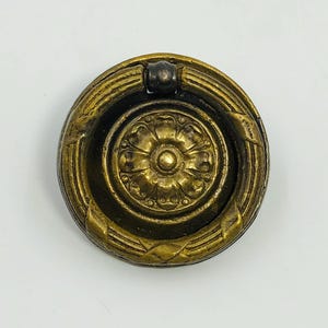 Può includere: Pomello per cassetto in ottone antico con design rotondo. Il pomello presenta un motivo floreale centrale circondato da anelli concentrici e dettagli decorativi. La maniglia è una piccola barra arcuata. L'hardware ha un aspetto vintage.