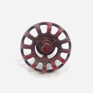 Puede incluir: Una manija de válvula circular de metal rojo oscuro que presenta una serie de radios arqueados espaciados uniformemente que irradian desde un cubo central. La superficie muestra signos de desgaste y antigüedad, con un acabado desgastado.
