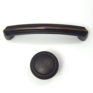 Pode incluir: Um conjunto de duas peças de ferragens de armário em bronze escuro. Uma é uma alça curva com um design canelado, e a outra é um botão redondo com um padrão circular. Ambas as peças têm um acabamento escovado.