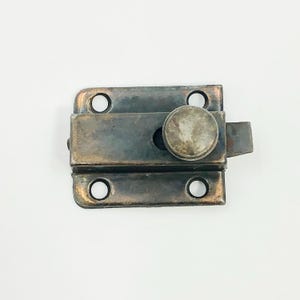 May include: A vintage metal latch with a tarnished finish. The latch features a rectangular base with rounded corners and four screw holes. A circular knob and a sliding bolt complete the design. Ideal for antique furniture or decorative projects.