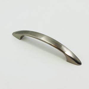May include: A curved cabinet handle with a smooth, brushed metallic finish. The hardware features a sleek, arched design with tapered ends, providing a functional grip for drawers or doors. The surface reflects light, highlighting the silver tone.