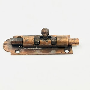 May include: A vintage copper-toned metal bolt latch with a ball-shaped handle. The latch has a rectangular base with rounded edges and two screw holes. The bolt is extended, showing signs of age and wear, suggesting it is an antique or reclaimed hardware.