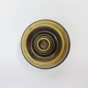 Op de afbeelding: Een vintage messing deurknop met een cirkelvormig ontwerp. De knop heeft concentrische cirkels en een gedetailleerd middenpatroon. Het messing heeft een warme, verouderde patina, met donkerdere tinten in de verzonken gebieden.