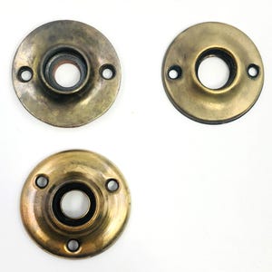 May include: Three antique brass door rosettes. Each circular rosette features a central hole and screw holes. The brass has a tarnished, aged appearance, with variations in colour. Ideal for restoration projects.
