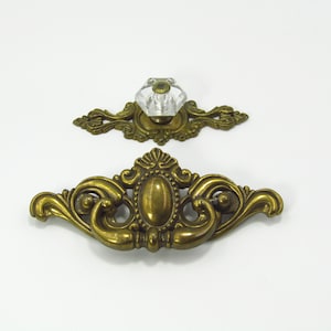 May include: Two antique brass drawer pulls. The top pull has a clear, faceted glass knob. The bottom pull is a larger, ornate brass handle with an oval centre. Both have a vintage, decorative design.