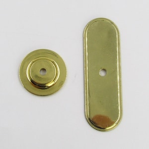 May include: Two brass-coloured hardware pieces: a round disc and a rectangular plate with rounded ends. Both have a small hole in the centre. These items are likely for furniture or cabinet construction.