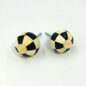 May include: Two spherical cabinet knobs featuring a checkered pattern of alternating black and off-white segments. Each knob has a protruding metal screw bolt extending from the back, designed for attachment to furniture or cabinetry doors.
