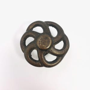 May include: A circular metal knob with an antique bronze finish, featuring a central solid disc surrounded by six symmetrical, curved, open-work petal shapes that create a continuous, flowing pattern across the face of the hardware piece.
