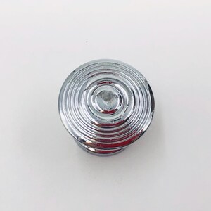 May include: A circular cabinet knob with a polished chrome finish, featuring a series of concentric, raised circular ridges that radiate from the center to the outer edge, creating a textured, stepped profile on the front-facing surface.