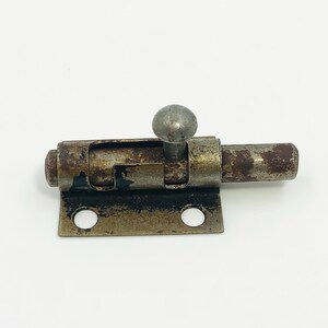 May include: A vintage metal bolt latch with a weathered, rusty appearance. The latch features a sliding bolt, a round knob, and a rectangular base with two circular holes. The metal has a mottled brown and gray color, suggesting age and use.