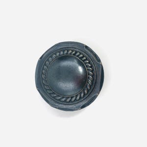 May include: A circular cabinet knob with a dark bronze finish. The knob features a smooth, slightly domed center surrounded by a raised, twisted rope-like border. The outer edge of the knob has a subtle, multi-faceted octagonal shape.
